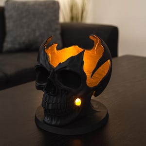 Könnte beinhalten: Eine schwarze, schädelartige Dekorationslampe mit einem leuchtend orangefarbenen Innenraum. Der Schädel weist detaillierte Merkmale auf und steht auf einem runden Sockel. Das Innere leuchtet und erzeugt einen dramatischen Effekt. Ein kleines Licht scheint aus der Wange des Schädels.