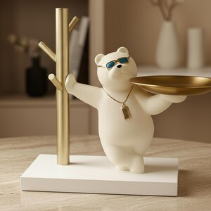 Könnte beinhalten: Eine weiße Bärenfigur mit Sonnenbrille und goldener Halskette, die ein goldfarbenes Tablett hält. Der Bär steht auf einem weißen Sockel, links daneben ein goldfarbener baumförmiger Kleiderständer. Die Figur ist ein Dekorationsartikel.