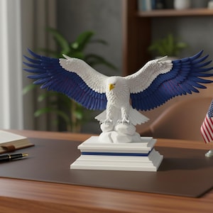 K&ouml;nnte beinhalten: Eine wei&szlig;e und blaue Adlerstatue mit ausgebreiteten Fl&uuml;geln, die auf einem wei&szlig;en Sockel mit blauen Akzenten sitzt. Der Adler hat einen gelben Schnabel und wird auf einem Schreibtisch mit einer kleinen amerikanischen Flagge ausgestellt.