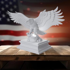 K&ouml;nnte beinhalten: Eine wei&szlig;e Skulptur eines Adlers mit ausgebreiteten Fl&uuml;geln, die auf einem rechteckigen Sockel steht. Der Adler ist detailliert mit sichtbaren Federn. Der Hintergrund zeigt eine verschwommene amerikanische Flagge und eine Holzoberfl&auml;che.