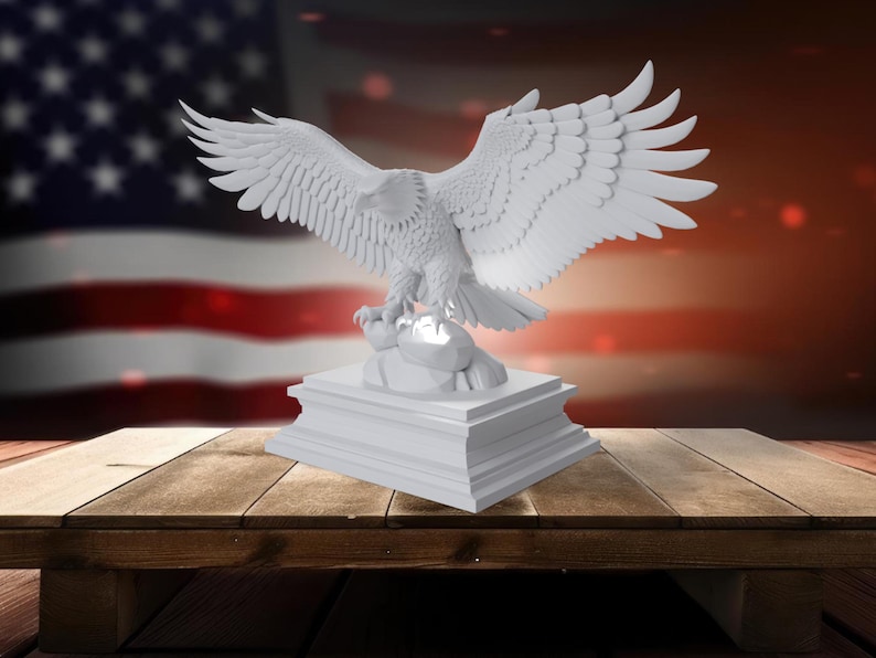 K&ouml;nnte beinhalten: Eine wei&szlig;e Skulptur eines Adlers mit ausgebreiteten Fl&uuml;geln, die auf einem rechteckigen Sockel steht. Der Adler ist detailliert mit sichtbaren Federn. Der Hintergrund zeigt eine verschwommene amerikanische Flagge und eine Holzoberfl&auml;che.