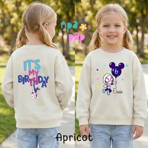 Op de afbeelding: Abrikooskleurige sweatshirt met een kleurrijke afbeelding van een superheld die een ballon met het nummer 6 vasthoudt. De tekst "Elodie" staat eronder. De achterkant van de trui heeft de tekst "It's My Birthday" met een superheldenafbeelding.