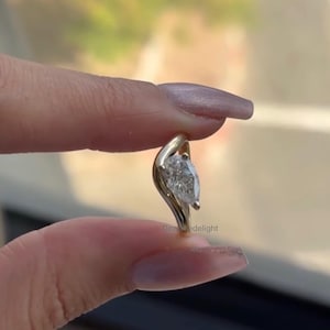 Puede incluir: Un colgante de oro con un diamante en forma de pera. El colgante tiene un diseño único y fluido. El diamante es claro y brillante. El texto "Dezadelight" es visible en la parte inferior derecha.