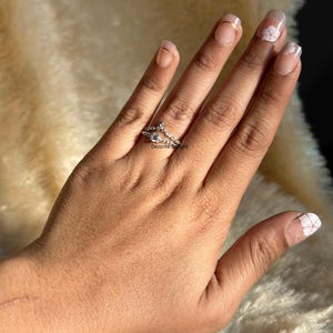 May include: A silver ring set with a small, clear gemstone and smaller accent stones. The ring has a unique, layered design with a chevron shape. The text "Dezzrite Delight" is visible on the ring.
