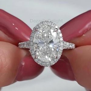 May include: A dazzling engagement ring featuring a large, oval-cut diamond surrounded by a halo of smaller stones. The band is also adorned with sparkling accents. The ring is held up by a person with pink nail polish.