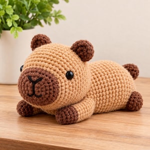 May include: A crocheted capybara plush toy featuring a light tan body with dark brown accents on the ears, snout, and feet. The stuffed animal has two round black plastic eyes and a stitched dark brown nose and mouth in a horizontal position.
