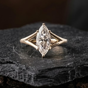 Puede incluir: Un anillo de oro con una piedra preciosa transparente de corte marquesa en un engaste de garras. La banda tiene un diseño de vástago dividido que se estrecha hacia la piedra central, creando un aspecto geométrico de oro amarillo pulido.