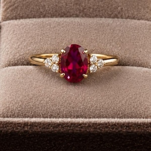 May include: A gold ring featuring a central oval-cut deep red gemstone held by four prongs, flanked by three small round white accent stones on each side, presented inside a brown velvet-lined jewellery box with a hinged lid and soft interior.