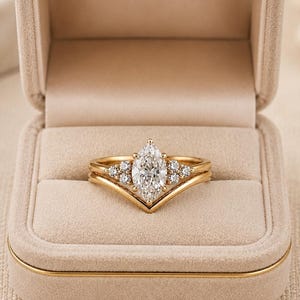 May include: A gold ring set featuring a central marquise-cut stone flanked by three smaller round stones on each side, nested within a V-shaped gold band. The jewellery is displayed inside a beige velvet ring box with a hinged lid and gold trim.