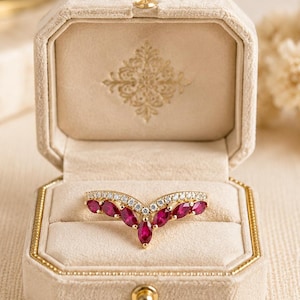 Könnte beinhalten: Ein goldener Ring mit V-förmigem Design, besetzt mit roten Edelsteinen und kleinen weißen Diamanten. Der Ring wird in einer offenen, beigefarbenen Samt-Schmuckschatulle mit goldenem Verschluss und Zierleiste präsentiert.