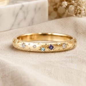 May include: A gold band ring featuring a series of engraved starburst patterns. The ring is set with multiple small round gemstones, including clear white stones, a light blue stone, and a deep purple stone, all embedded flush into the metal surface.