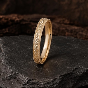 May include: A gold ring featuring an intricate, embossed floral scroll pattern along the outer band, bordered by delicate beaded edges. The metal has a polished, reflective finish, highlighting the detailed relief work on the surface of the band.