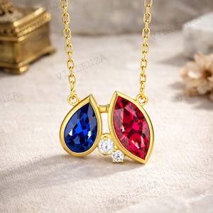 Op de afbeelding: Een gouden ketting met een hanger. De hanger heeft een blauwe, druppelvormige edelsteen en een rode, bladvormige edelsteen. Twee kleine witte edelstenen zijn tussen de grotere stenen gezet. De ketting zit aan een gouden ketting.