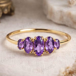 Könnte beinhalten: Ein goldener Ring mit fünf ovalen, lila Edelsteinen. Die Edelsteine sind in einer Reihe über der Oberseite des Rings angeordnet. Der Ring hat ein dünnes Band. Der Ring wird auf einer weißen Oberfläche präsentiert.