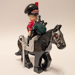 May include: A Lego figure of a Scottish bagpiper in a red jacket and black hat, riding a black horse. The horse has a white saddle and a silver sword. The bagpiper is holding a bagpipe with a green and blue plaid pattern.
