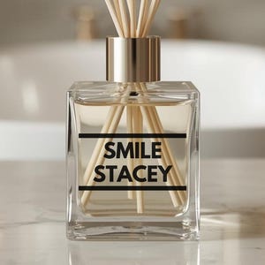 以下が含まれることがあります： 金色の襟と木のリードが付いた透明なガラスディフューザーボトル。「SMILE STACEY」の文字が黒色で書かれています。ディフューザーはバスルームの環境にある大理石の表面にあります。