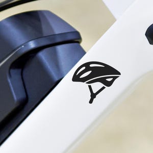 May include: A black helmet sticker on a white bicycle frame. The sticker depicts a stylized bicycle helmet, suggesting safety gear for cycling. The frame also features a dark blue component, possibly a battery or motor.