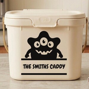 May include: A cream-coloured caddy with a lid and a handle. A black cartoon monster graphic with three eyes and a wide grin is on the front. The text "THE SMITHS CADDY" is below the graphic in a black font.