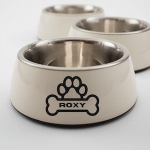 May include: Three cream-coloured pet bowls with stainless steel interiors. The front bowl has a black paw print and bone design with the name "ROXY". These bowls are suitable for pet food or water.