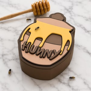 Puede incluir: Un tarro de miel marrón con tapa color canela y un diseño de goteo de miel amarillo que presenta la palabra HUNNY en letras marrones en relieve. El tarro se acompaña de un cucharón de miel de madera y tres pequeñas figuras de abejas.