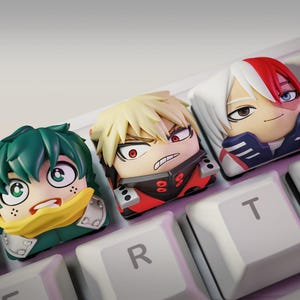 Puede incluir: Tres teclas artesanales personalizadas con forma de cabezas de personajes de anime con cabello verde, rubio y cabello dividido en blanco y rojo, instaladas en un teclado mecánico blanco con las letras R y T visibles en las teclas adyacentes.