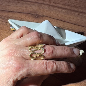Könnte beinhalten: Ein goldfarbener Ring mit einem wellenförmigen Design. Der Ring wird an einem Finger getragen, im Hintergrund befinden sich ein weißes Papierboot und eine Holzoberfläche. Das Design des Rings besteht aus einer Reihe verbundener Kurven.