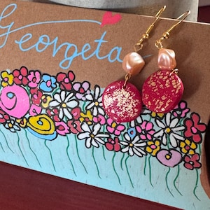Könnte beinhalten: Goldfarbene Ohrringe mit roten, goldfarben gefleckten, kreisförmigen Anhängern und pfirsichfarbenen Perlen. Die Ohrringe hängen vor einer floralen Illustration mit dem Namen "Georgeta" in Schreibschrift. Die Illustration zeigt rosa, weiße, blaue und gelbe Blumen.