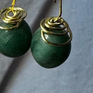 Könnte beinhalten: Ein Paar Ohrhänger mit dunkelgrünen, ovalen Steinen. Jeder Stein hat eine goldfarbene Drahtspirale und einen goldfarbenen Haken. Die Ohrringe werden vor einem gedämpften grauen Hintergrund gezeigt.