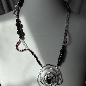 Könnte beinhalten: Eine handgefertigte Halskette mit einem silberfarbenen, spiralförmigen Anhänger und einem schwarzen Mittelteil. Die Halskette enthält schwarze, klare und burgunderfarbene Perlen sowie schwarze zylindrische Akzente. Die Halskette wird auf einer weißen Schaufensterpuppe präsentiert.