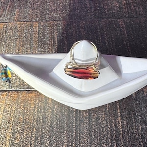 Könnte beinhalten: Ein goldfarbener Ring mit einem rechteckigen, rotbraunen Stein. Der Ring liegt auf einer weißen, bootsförmigen Schale vor einem dunklen Holzmaserungshintergrund. Das Design des Rings umfasst einen Band und einen rechteckigen Stein.