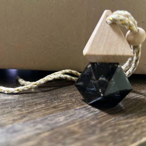 Könnte beinhalten: Ein schwarzer, facettierter Kristallanhänger mit einem dreieckigen Holzaufsatz und einem beige-goldenen Seil. Der Anhänger hängt an einer Holzoberfläche. Der Kristall hat eine dunkle, reflektierende Farbe.