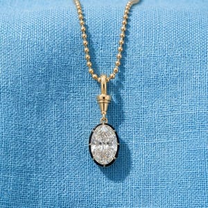 May include: A gold-toned ball chain necklace featuring a pendant with a central oval-shaped clear stone surrounded by a thin black border. The pendant is suspended from a gold-toned conical bail that connects directly to the delicate beaded chain.