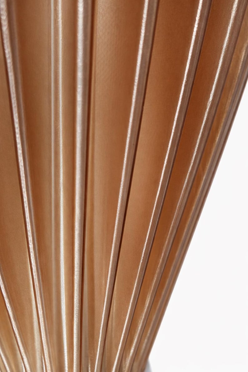 Pode incluir: Close-up de um tecido plissado em um tom marrom claro e quente. O tecido tem dobras verticais com destaques sutis, criando um padr&atilde;o texturizado em forma de leque. O fundo &eacute; branco.