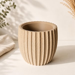 Könnte beinhalten: Ein zylindrischer Pflanztopf mit einer texturierten, gerippten Außenseite in hellem Beige. Das Gefäß verfügt über einen glatten, flachen Rand und einen tiefen, hohlen Innenraum, der ein durchgehendes vertikales Rillenmuster auf der Oberfläche zeigt.