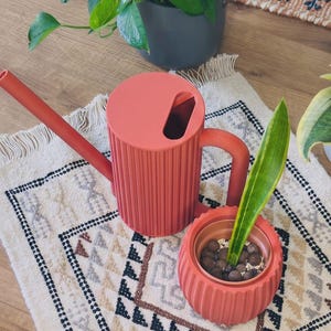 May include: A terracotta-coloured cylindrical watering can with a long spout and a ribbed texture, paired with a matching ribbed planter containing a single green and yellow variegated leaf planted in dark brown clay pebbles.
