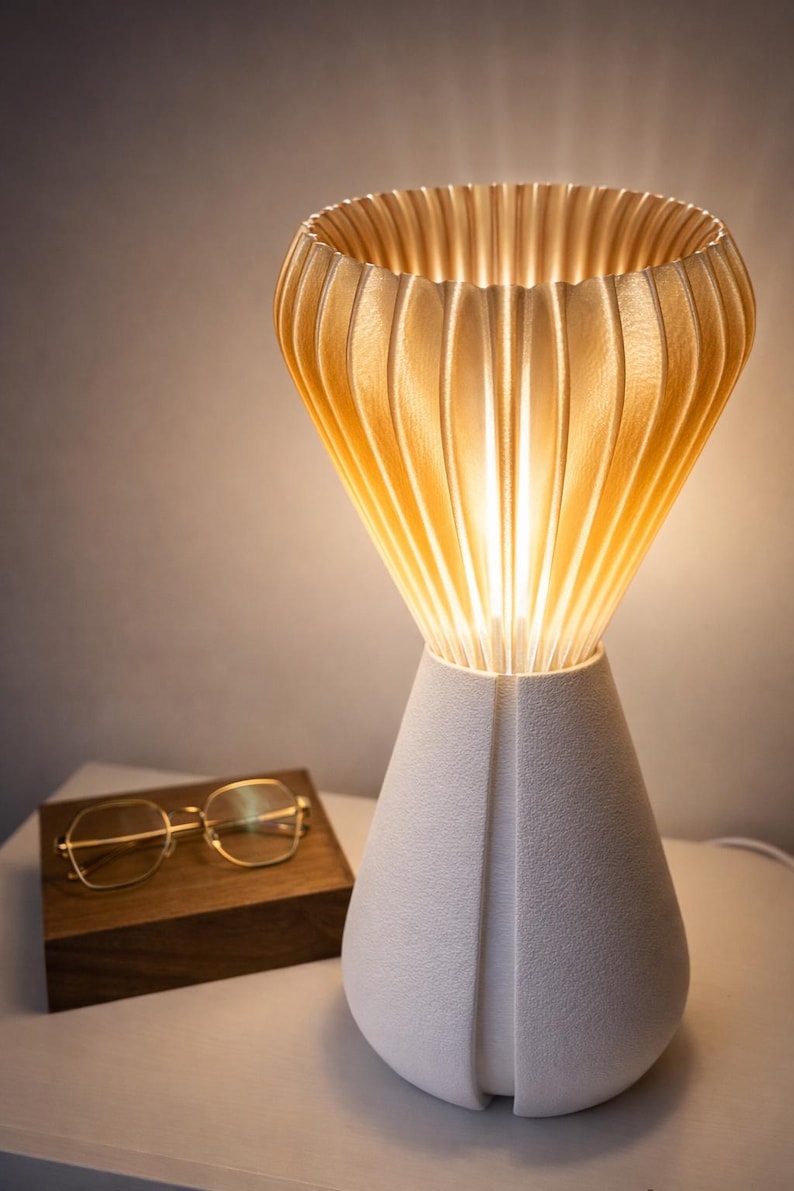 Pode incluir: Uma moderna lumin&aacute;ria de mesa com base branca e abajur texturizado de cor clara. O abajur tem um design canelado e a lumin&aacute;ria est&aacute; acesa. &Oacute;culos com arma&ccedil;&atilde;o dourada sobre uma caixa de madeira. Decora&ccedil;&atilde;o de casa.