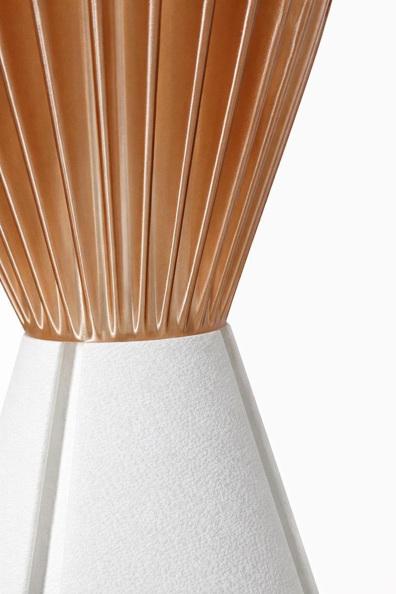 Pode incluir: Close-up de uma l&acirc;mpada moderna. A parte superior &eacute; dourada met&aacute;lica quente com ranhuras verticais. A base &eacute; branca texturizada, com formato geom&eacute;trico. A l&acirc;mpada est&aacute; em um fundo branco.