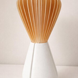 Pode incluir: Um vaso decorativo moderno com uma base geom&eacute;trica branca e um topo em forma de leque feito de ripas de madeira verticais castanho-claro. O vaso tem um design &uacute;nico e contempor&acirc;neo.