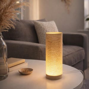 May include: A cylindrical table lamp with a textured, woven shade emits a warm glow. The lamp sits on a white table, next to a small wooden dish with gold rings. A sofa and decorative dried plants are in the background.