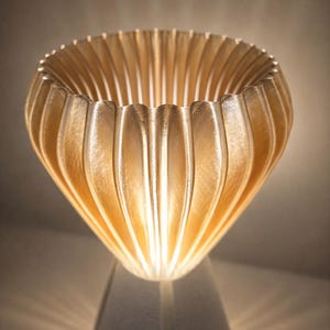 Pode incluir: Uma l&acirc;mpada decorativa com uma sombra dourada e canelada e uma base branca. A sombra tem um design texturizado &uacute;nico, e a luz brilha atrav&eacute;s, criando um brilho quente. O design da l&acirc;mpada &eacute; moderno e art&iacute;stico.