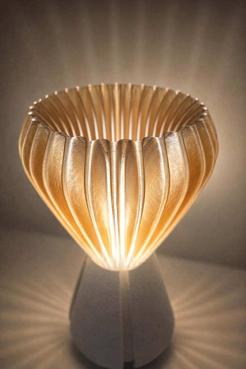 Pode incluir: Uma l&acirc;mpada decorativa com uma sombra dourada e canelada e uma base branca. A sombra tem um design texturizado &uacute;nico, e a luz brilha atrav&eacute;s, criando um brilho quente. O design da l&acirc;mpada &eacute; moderno e art&iacute;stico.