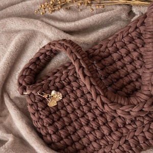 May include: A brown, hand-knitted tote bag with a braided handle and a decorative charm. The bag is made of thick, soft yarn and has a textured appearance. The bag is resting on a soft, brown blanket.