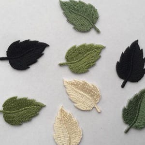 Op de afbeelding: Een verzameling van acht geborduurde bladpatches in verschillende kleuren, waaronder donkergroen, lichtgroen, zwart en crème. Elke patch heeft een gedetailleerde stiktextuur met een centrale nerf en gekartelde randen, verspreid gerangschikt.