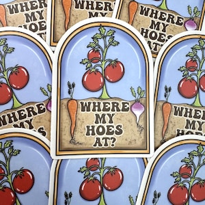 May include: A rectangular sticker with rounded top corners featuring a garden scene with red tomatoes on a vine, a carrot, and a turnip in soil. The text WHERE MY HOES AT? is printed in the center. The background is light blue with a tan soil area.