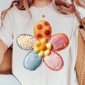May include: White t-shirt featuring a large, colorful flower applique. The flower has petals in orange, pink, blue, and white, with yellow and orange polka dots. The center of the flower is yellow with a textured design.