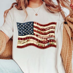 Puede incluir: Una camiseta blanca con un diseño desgastado de la bandera estadounidense. La bandera tiene un campo de estrellas azul y rayas rojas y blancas con patrones florales y de lunares. La bandera está cosida a la camiseta.