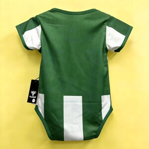 May include: Green baby bodysuit with white accents on the sleeves and lower back. The bodysuit has a ribbed texture and a tag with the brand name "hummel". The background is a solid yellow.