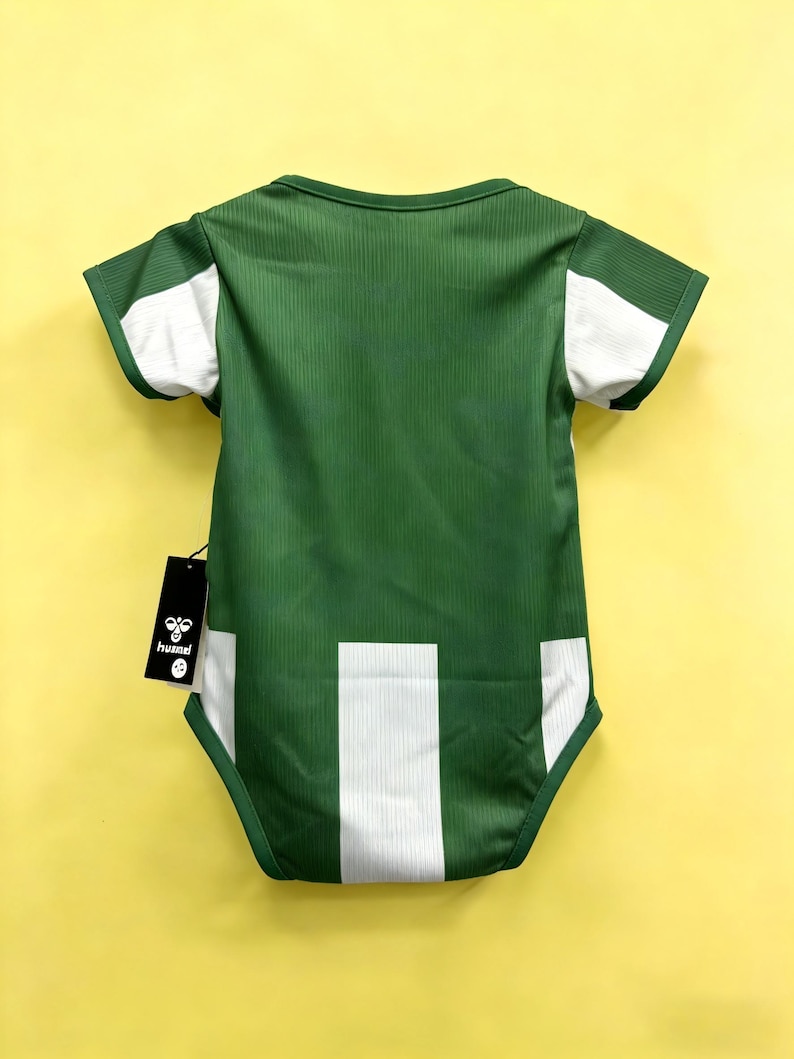 May include: Green baby bodysuit with white accents on the sleeves and lower back. The bodysuit has a ribbed texture and a tag with the brand name "hummel". The background is a solid yellow.