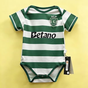 Puede incluir: Un body de bebé a rayas verdes y blancas con mangas cortas. El body presenta el texto "Betano" en negro y un escudo del equipo. El body tiene una etiqueta adjunta.