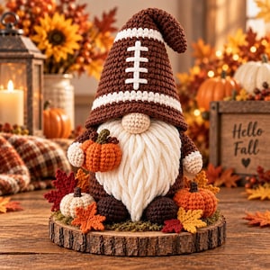 May include: A crochet gnome figurine featuring a brown and white football-patterned hat, a long white yarn beard, and a round nose. The gnome holds a small orange pumpkin and sits on a wooden base decorated with crochet pumpkins and autumn leaves.
